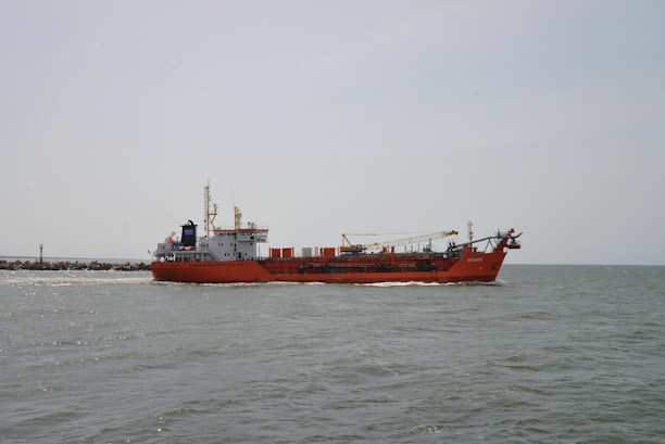 A large red cargo ship is sailing on a calm sea. The ship has various equipment and structures on its deck, including a crane. The horizon is visible in the background under a clear, pale sky.