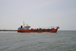 A large red cargo ship is sailing on a calm sea. The ship has various equipment and structures on its deck, including a crane. The horizon is visible in the background under a clear, pale sky.