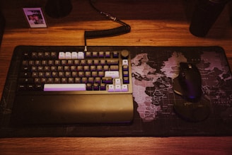 A mechanical keyboard with various colored keys rests on a world map-themed desk mat. Next to the keyboard is a black ergonomic mouse. A small photo in a frame is placed in the top left corner.