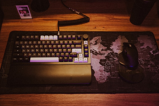 A mechanical keyboard with various colored keys rests on a world map-themed desk mat. Next to the keyboard is a black ergonomic mouse. A small photo in a frame is placed in the top left corner.