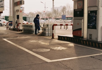 A person dressed in blue work attire is handling equipment near a row of large containers, possibly at a gas station. Multiple fuel pumps with signage and bollards are visible around the area. The background features parked cars and urban infrastructure such as streetlights and buildings.