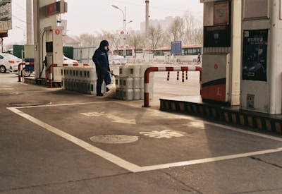 A person dressed in blue work attire is handling equipment near a row of large containers, possibly at a gas station. Multiple fuel pumps with signage and bollards are visible around the area. The background features parked cars and urban infrastructure such as streetlights and buildings.