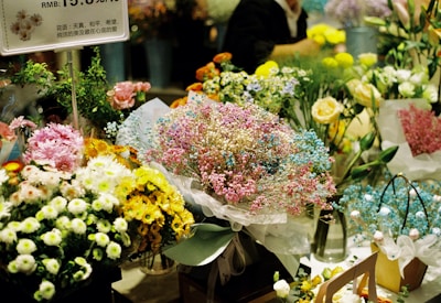 A vibrant bouquet shop featuring an array of colorful flowers, including chrysanthemums, lilies, and baby's breath. Bouquets are arranged neatly, displaying hues of pink, blue, yellow, and white. A small sign with Chinese characters and pricing information is visible. The setting is bustling and lively, with numerous arrangements ready for purchase.