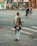 A person wearing casual clothing stands on a cobblestone path, gazing towards cyclists and motorcyclists on a street. The individual carries a camera and a bag, suggesting they might be a photographer. The background features urban buildings and trees.