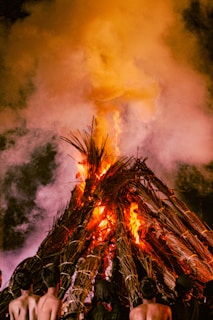 A large bonfire made of stacked wood and palm leaves is burning intensely, with flames rising high and smoke billowing into the air. The scene is likely taking place at night, and a few people are standing near the fire, partially visible from behind.