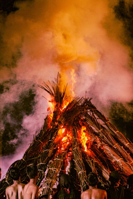 A large bonfire made of stacked wood and palm leaves is burning intensely, with flames rising high and smoke billowing into the air. The scene is likely taking place at night, and a few people are standing near the fire, partially visible from behind.