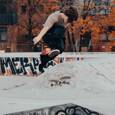 A skateboarder performs a mid-air trick against an urban backdrop with graffiti and autumn-colored trees. The skateboard is captured beneath the individual, emphasizing the dynamic nature of the stunt.