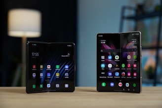 Two foldable smartphones are displayed on a wooden surface, partially unfolded with their screens showing various app icons. The background is softly blurred, featuring a lamp and some shelves.