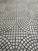 The image displays a tiled floor with a geometric pattern consisting of circular and square shapes. The tiles are arranged in concentric circles with small square tiles in varying shades of gray, creating a repetitive, symmetrical design.