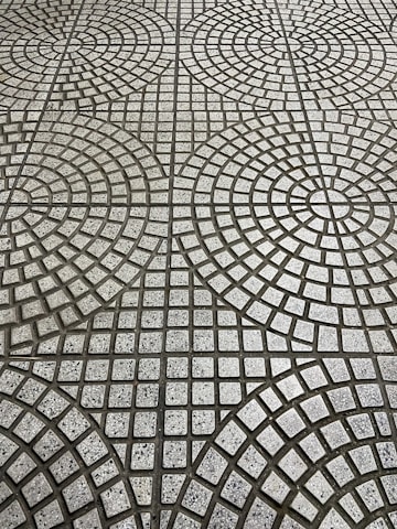 The image displays a tiled floor with a geometric pattern consisting of circular and square shapes. The tiles are arranged in concentric circles with small square tiles in varying shades of gray, creating a repetitive, symmetrical design.