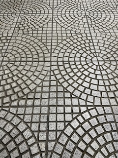 The image displays a tiled floor with a geometric pattern consisting of circular and square shapes. The tiles are arranged in concentric circles with small square tiles in varying shades of gray, creating a repetitive, symmetrical design.