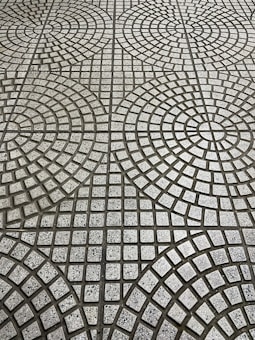 The image displays a tiled floor with a geometric pattern consisting of circular and square shapes. The tiles are arranged in concentric circles with small square tiles in varying shades of gray, creating a repetitive, symmetrical design.