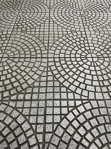 The image displays a tiled floor with a geometric pattern consisting of circular and square shapes. The tiles are arranged in concentric circles with small square tiles in varying shades of gray, creating a repetitive, symmetrical design.