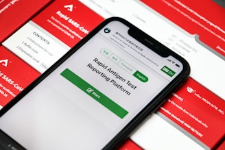 A smartphone displaying a rapid antigen test reporting platform application is placed on top of several red and white boxes labeled with 'Rapid SARS-CoV-2 Antigen Test.' The phone screen shows options for different languages and a start button for the application.