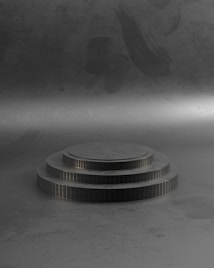 A three-tiered, round platform stands in a dimly lit room. Each tier is slightly recessed from the next and they are uniform in their grayscale color. The surrounding area has a textured, gradient backdrop with subtle streaks and shadows.