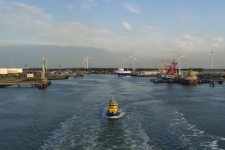 A harbor scene with a small yellow vessel navigating through a wide water channel surrounded by industrial infrastructure. In the background, there are several wind turbines, cranes, and larger ships docked along the piers. The sky is mostly clear with scattered clouds, and the sun casts a warm light over the scene.