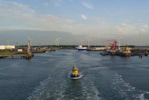 A harbor scene with a small yellow vessel navigating through a wide water channel surrounded by industrial infrastructure. In the background, there are several wind turbines, cranes, and larger ships docked along the piers. The sky is mostly clear with scattered clouds, and the sun casts a warm light over the scene.