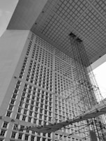 An upward perspective of a large, modern architectural structure featuring a geometric design with a grid-like pattern on the facade. The structure includes a massive overhanging roof, supported by intricate scaffolding or steel framework visible in the foreground.