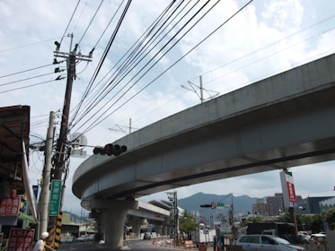 A large, elevated concrete highway or railway runs across the image, supported by sturdy pillars. Beneath it, a busy street scene includes power lines, traffic signals, and several vehicles. Buildings are visible in the background, with mountains in the distance under a partly cloudy sky.