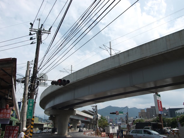A large, elevated concrete highway or railway runs across the image, supported by sturdy pillars. Beneath it, a busy street scene includes power lines, traffic signals, and several vehicles. Buildings are visible in the background, with mountains in the distance under a partly cloudy sky.