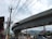 A large, elevated concrete highway or railway runs across the image, supported by sturdy pillars. Beneath it, a busy street scene includes power lines, traffic signals, and several vehicles. Buildings are visible in the background, with mountains in the distance under a partly cloudy sky.