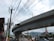 A large, elevated concrete highway or railway runs across the image, supported by sturdy pillars. Beneath it, a busy street scene includes power lines, traffic signals, and several vehicles. Buildings are visible in the background, with mountains in the distance under a partly cloudy sky.