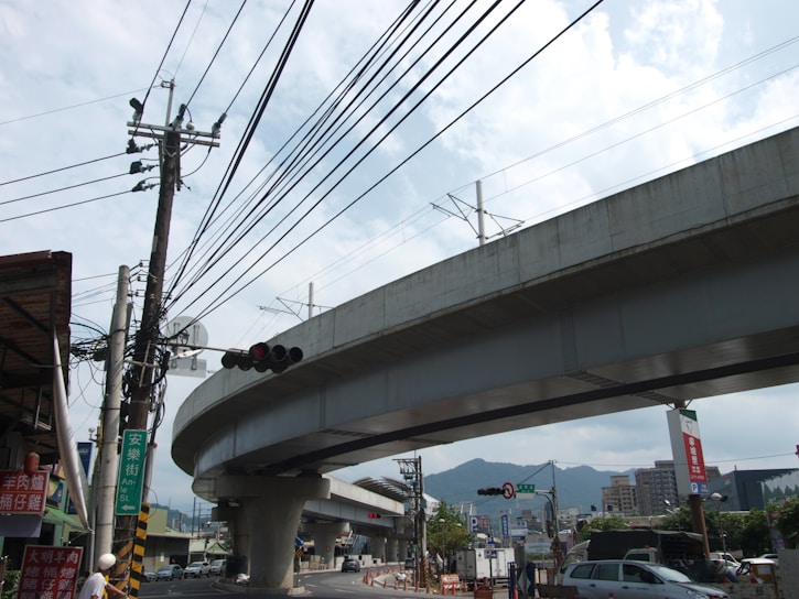 A large, elevated concrete highway or railway runs across the image, supported by sturdy pillars. Beneath it, a busy street scene includes power lines, traffic signals, and several vehicles. Buildings are visible in the background, with mountains in the distance under a partly cloudy sky.