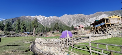 A picturesque mountainous landscape with tall, snow-capped peaks in the background. A lush green field and scattered wooden structures are in the foreground. A prominent purple tent and a wooden building with the sign 'Hotel' are visible, surrounded by rustic wooden fences. Tall evergreen trees can be seen throughout the scene.