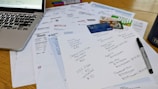 A cluttered workspace featuring a laptop, various bills, a list of subscriptions, credit cards, and a marker. The papers include invoices from different utility companies and entertainment services, with one paper having handwritten notes detailing subscription plans and prices.