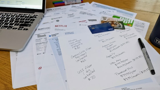 A cluttered workspace featuring a laptop, various bills, a list of subscriptions, credit cards, and a marker. The papers include invoices from different utility companies and entertainment services, with one paper having handwritten notes detailing subscription plans and prices.