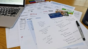 A cluttered workspace featuring a laptop, various bills, a list of subscriptions, credit cards, and a marker. The papers include invoices from different utility companies and entertainment services, with one paper having handwritten notes detailing subscription plans and prices.