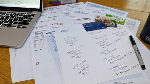 A cluttered workspace featuring a laptop, various bills, a list of subscriptions, credit cards, and a marker. The papers include invoices from different utility companies and entertainment services, with one paper having handwritten notes detailing subscription plans and prices.