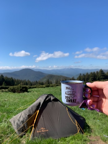 A hand holds a metal mug with an inspirational quote written on it, set against a backdrop of a lush green meadow, a pitched tent, and distant mountains under a clear blue sky.