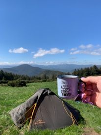 A hand holds a metal mug with an inspirational quote written on it, set against a backdrop of a lush green meadow, a pitched tent, and distant mountains under a clear blue sky.