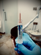 A gloved hand holds a syringe labeled 'WHITER IMAGE' containing a gel used for teeth whitening. The background shows a dental clinic setting with a dental chair and equipment.