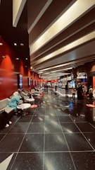 A modern cinema lobby with a sleek design featuring red walls and illuminated ceilings. People are seated on gray couches along the left side, engaging in conversations or using their phones. Posters line the walls, and the floor is polished black with reflective surfaces. Towards the back, there is a counter with a 'VIP Cashier' sign, and a few people are waiting in line.