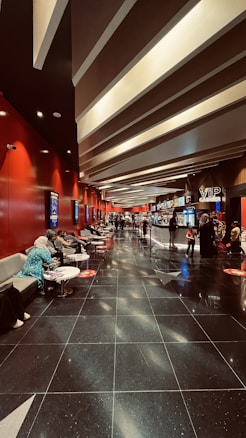 A modern cinema lobby with a sleek design featuring red walls and illuminated ceilings. People are seated on gray couches along the left side, engaging in conversations or using their phones. Posters line the walls, and the floor is polished black with reflective surfaces. Towards the back, there is a counter with a 'VIP Cashier' sign, and a few people are waiting in line.