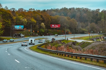 A highway scene featuring multiple vehicles, including cars and a large truck, traveling on a divided road. The road curves with a metal guardrail along the edge. On either side of the highway, there are billboards and traffic lights. The background includes a forested area with trees in various stages of foliage and a cloudy sky above.