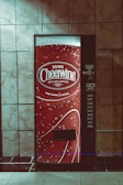 A vending machine with a bright red design featuring white text and decorative bubbles. It has the branding 'Drink Cheerwine Bottling Company' prominently displayed. The machine is set against a tiled wall in a dimly lit area.