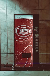 A vending machine with a bright red design featuring white text and decorative bubbles. It has the branding 'Drink Cheerwine Bottling Company' prominently displayed. The machine is set against a tiled wall in a dimly lit area.
