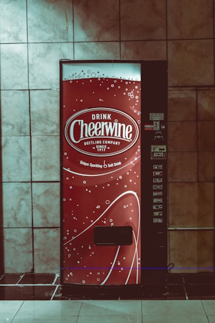 A vending machine with a bright red design featuring white text and decorative bubbles. It has the branding 'Drink Cheerwine Bottling Company' prominently displayed. The machine is set against a tiled wall in a dimly lit area.