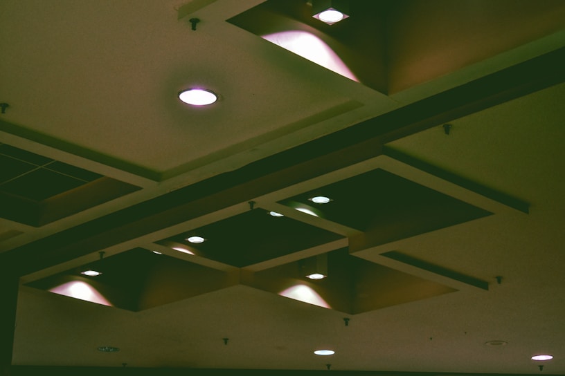 A ceiling featuring multiple recessed lighting fixtures, some emitting warm light. The design incorporates square and rectangular elements, creating a geometric pattern that adds depth and contrast. Shadows and highlights interplay across the surfaces.
