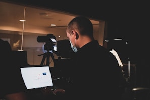 A person wearing a face mask is working in a dimly lit room, appearing focused on a laptop. A video camera is mounted on a tripod pointing towards a window, indicating some form of recording setup. The room has a professional and technical ambiance, possibly a studio or control room.