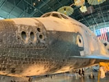 A close-up view of a space shuttle on display in a museum. The shuttle is large and partially black with thermal tiles covering its underside. The name 'Discovery' is visible on the side. Overhead, there is a display of satellite models and American flags in the background. The interior of the museum is spacious with a high ceiling.