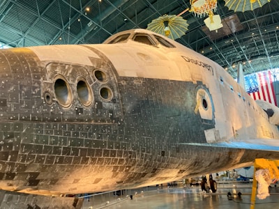 A close-up view of a space shuttle on display in a museum. The shuttle is large and partially black with thermal tiles covering its underside. The name 'Discovery' is visible on the side. Overhead, there is a display of satellite models and American flags in the background. The interior of the museum is spacious with a high ceiling.