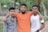 Three young men stand closely together, with the man in the center draping his arms over the shoulders of the others. The background features lush greenery and a stone path.