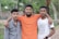 Three young men stand closely together, with the man in the center draping his arms over the shoulders of the others. The background features lush greenery and a stone path.