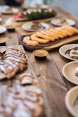 A variety of sliced fruits and desserts are arranged on wooden platters and plates on a rustic wooden table. The foreground features a platter of sliced pastries with a drizzle of chocolate or caramel, while the background shows sliced cantaloupe and what appears to be a smaller plate of desserts. The setup has a casual yet elegant presentation typical of a buffet or catered event.