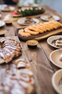 A variety of sliced fruits and desserts are arranged on wooden platters and plates on a rustic wooden table. The foreground features a platter of sliced pastries with a drizzle of chocolate or caramel, while the background shows sliced cantaloupe and what appears to be a smaller plate of desserts. The setup has a casual yet elegant presentation typical of a buffet or catered event.