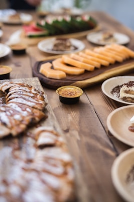 A variety of sliced fruits and desserts are arranged on wooden platters and plates on a rustic wooden table. The foreground features a platter of sliced pastries with a drizzle of chocolate or caramel, while the background shows sliced cantaloupe and what appears to be a smaller plate of desserts. The setup has a casual yet elegant presentation typical of a buffet or catered event.